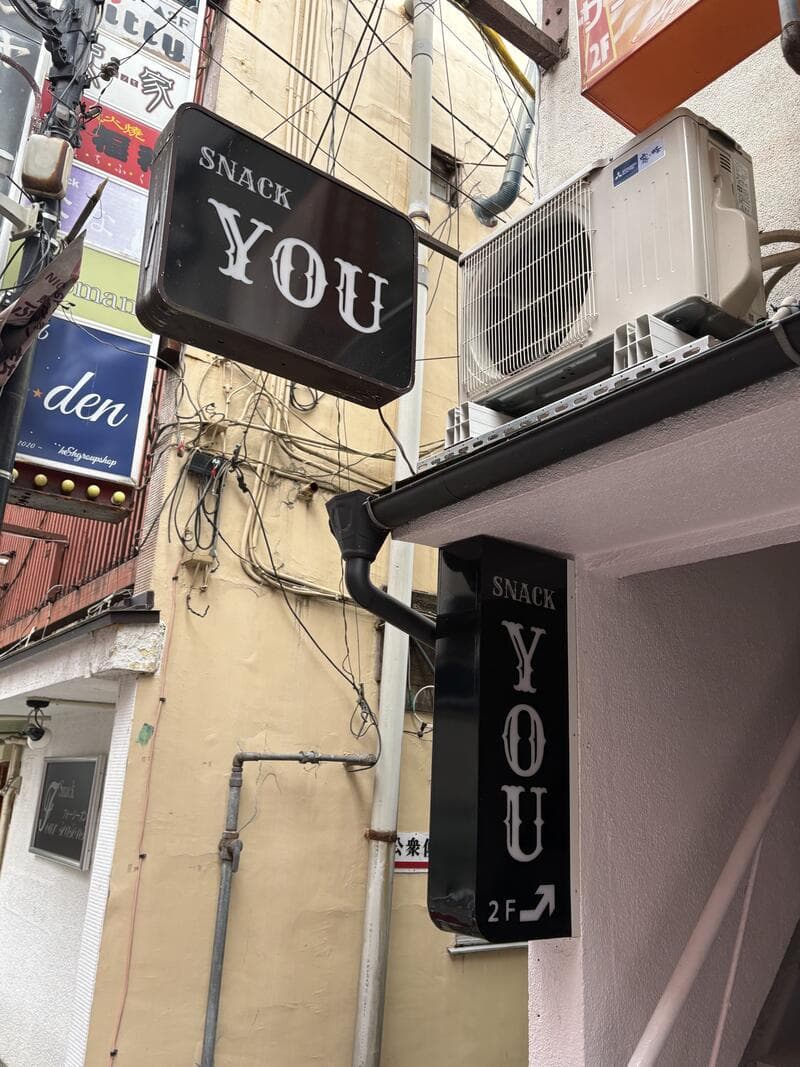 黒い箱看板に白文字を置いた「SNACK YOU」の2階案内