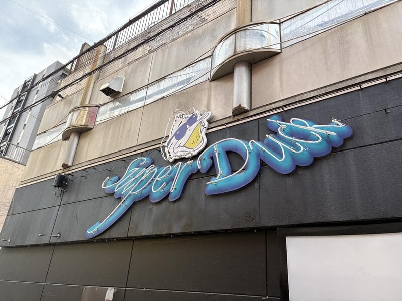 黒いファサードに青い筆記体ロゴを置いた「Super Duck」看板